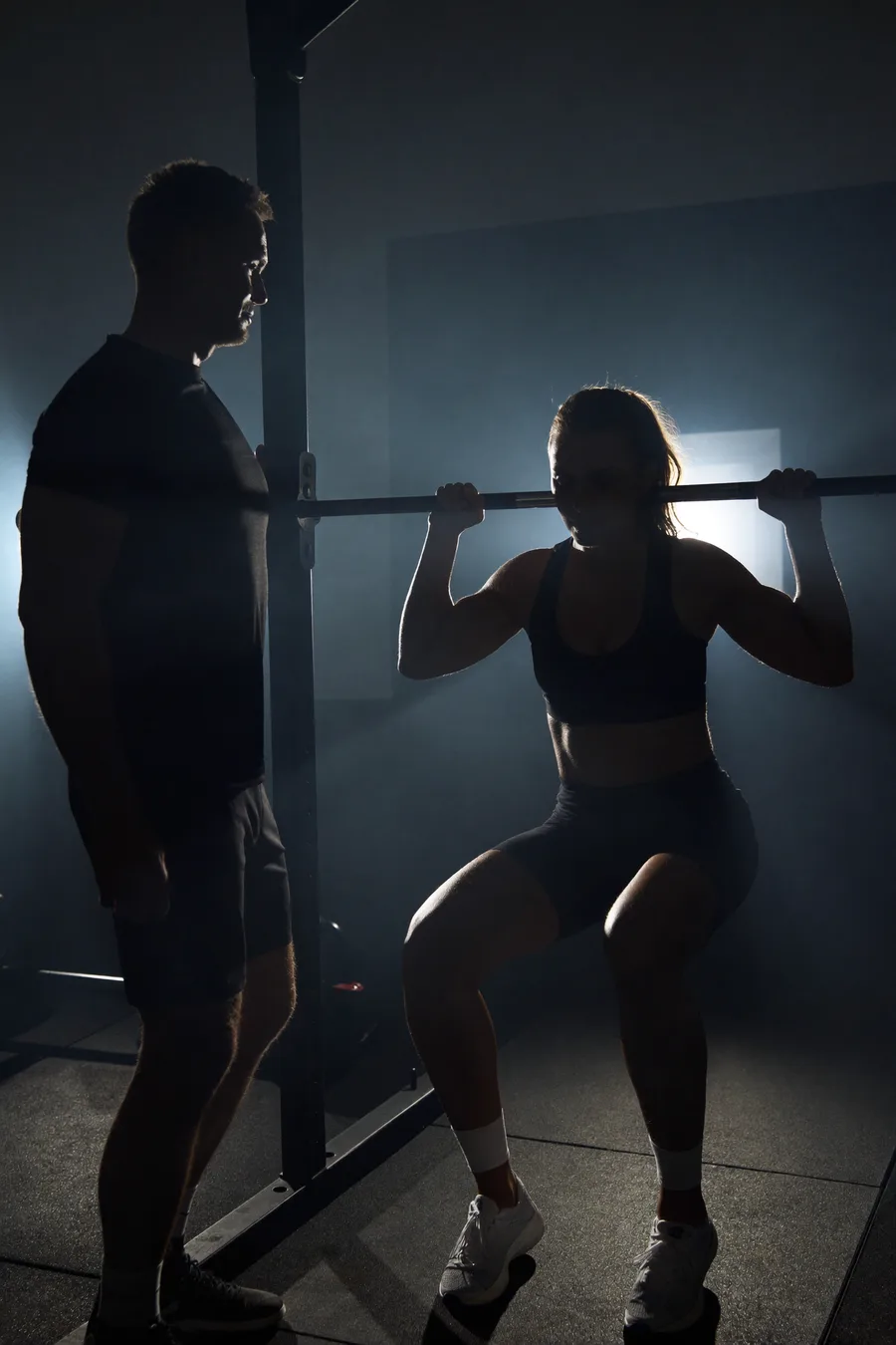 Coach sichert Athleten am Rack — dramatisches Gegenlicht im dunklen Studio